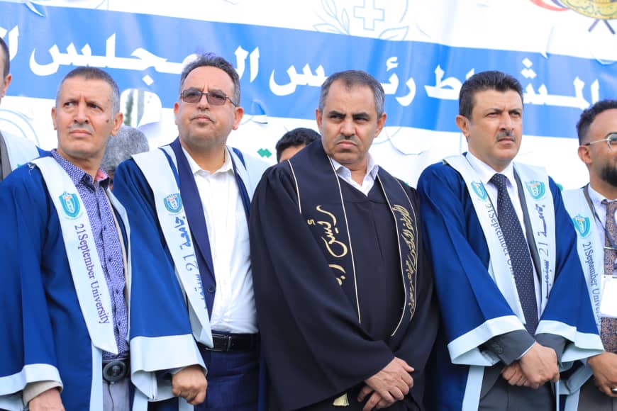 1003982242 - جامعة 21 سبتمبر Group of men in blue graduation gowns posing for a photo at a formal ceremony, with a blue banner in Arabic behind them.