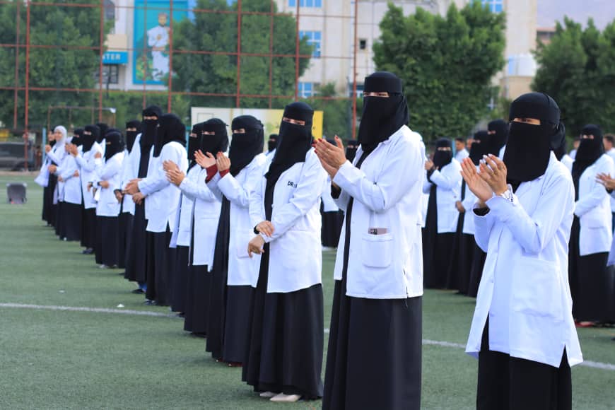 1003982245 - جامعة 21 سبتمبر Group of women in white lab coats and black hijabs standing in a line on a grassy field, clapping at an outdoor ceremony.