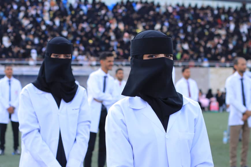 1003982248 - جامعة 21 سبتمبر Two women in white lab coats and black niqabs stand on a sports field as a large crowd watches.