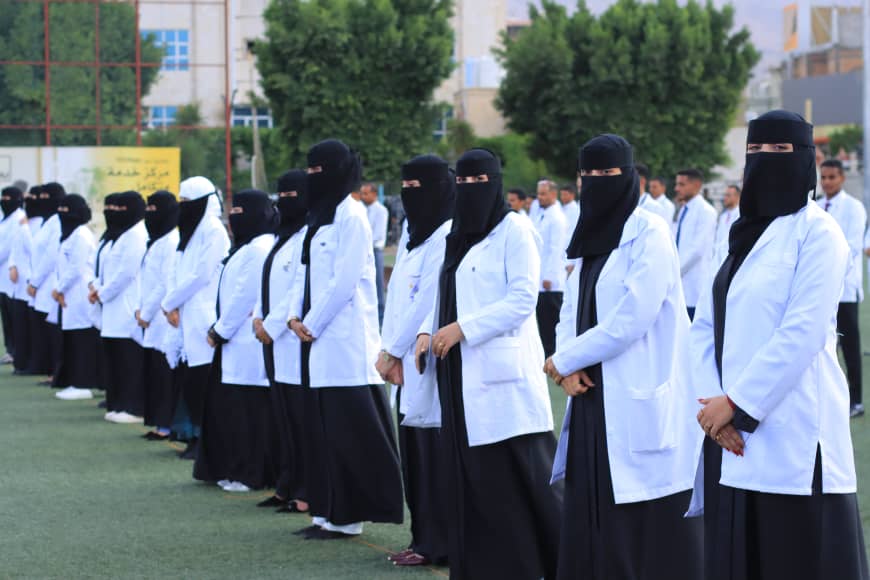 1003982251 - جامعة 21 سبتمبر Group of women in white lab coats and black veils standing in a line on an outdoor field, facing forward.