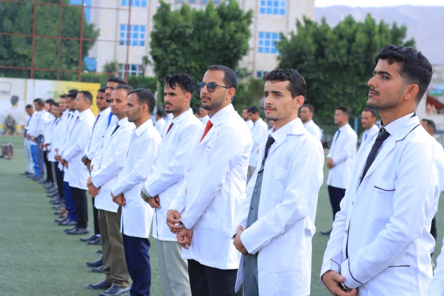 1003982254 - جامعة 21 سبتمبر Line of men in white lab coats and ties standing in a row during an outdoor event on a sports field with buildings and trees in the background.