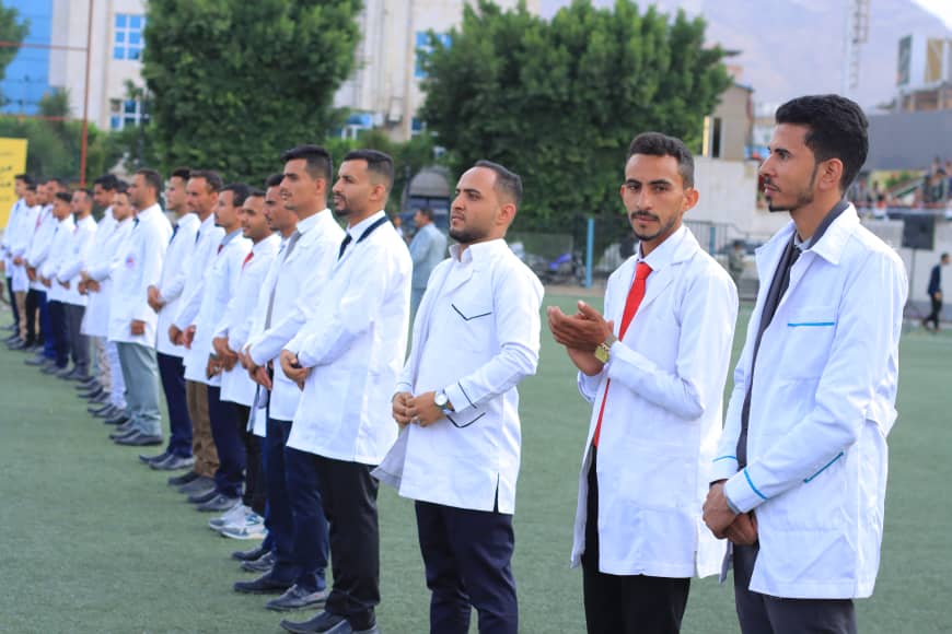 1003982257 - جامعة 21 سبتمبر Group of men in white lab coats standing in a line on a sports field, facing forward.