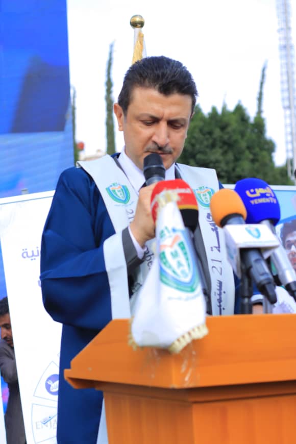 1003982260 - جامعة 21 سبتمبر Man in a blue academic gown and sash delivering a speech at a podium with multiple microphones