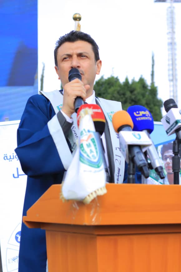 1003982263 - جامعة 21 سبتمبر Man in a blue robe speaks into a microphone at a wooden podium during a press conference, surrounded by multiple colorful microphones.