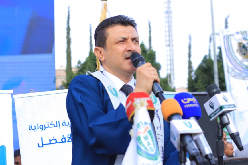 1003982266 - جامعة 21 سبتمبر Man in a blue suit speaks into a microphone at a press conference, surrounded by multiple news microphones.
