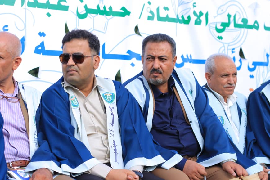 1003982269 - جامعة 21 سبتمبر Group of men in blue and white ceremonial robes seated at an event, with a banner containing Arabic text behind them.