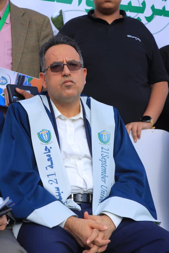 1003982272 - جامعة 21 سبتمبر Man wearing sunglasses, white shirt, and blue ceremonial robe with a sash bearing a crest and the text '21 September' at an event.