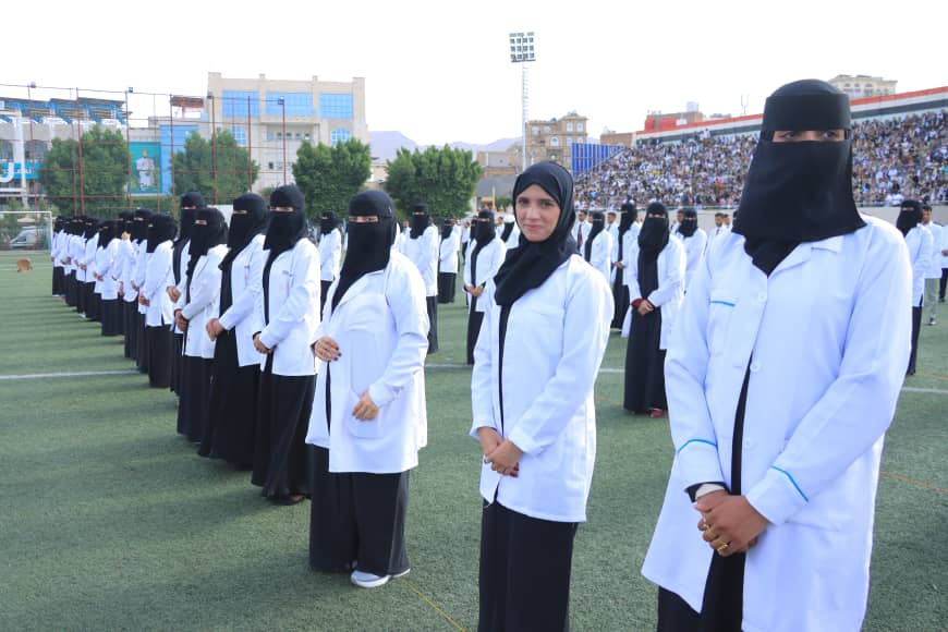 1003982275 - جامعة 21 سبتمبر Line of women in white lab coats and black hijabs standing in formation on a sports field during a ceremony at a stadium stead.
