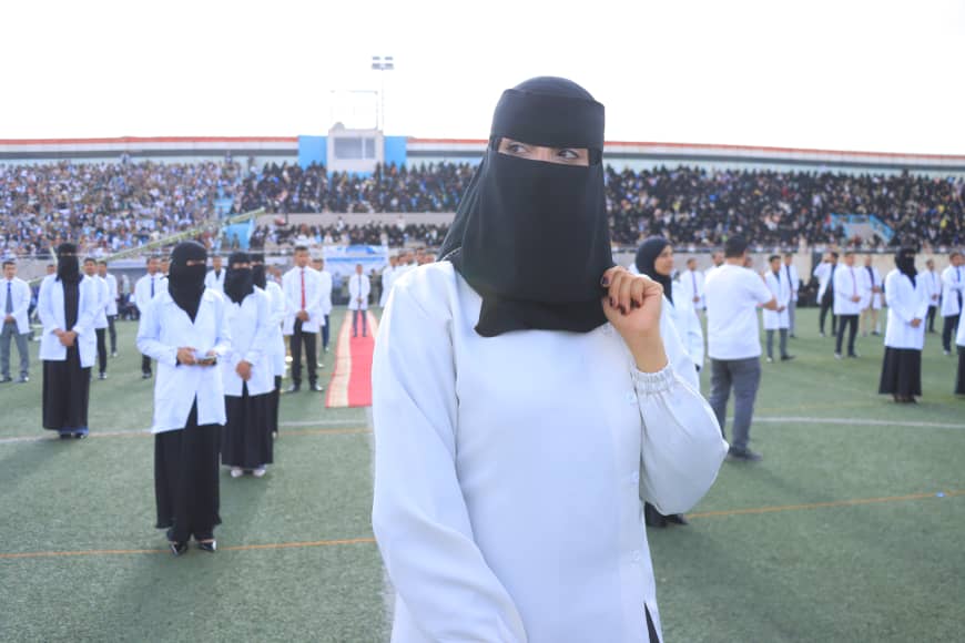 1003982278 - جامعة 21 سبتمبر Woman wearing a black niqab stands at the front on a football field during a large group event, with many students in white shirts in the background.