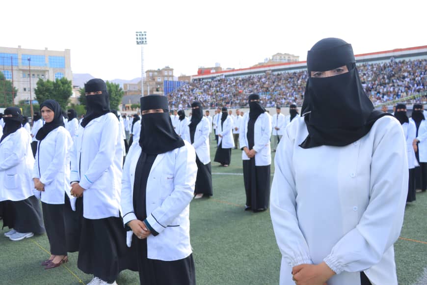 1003982281 - جامعة 21 سبتمبر Group of women in white lab coats and black hijabs standing on a stadium field during a large outdoor ceremony.