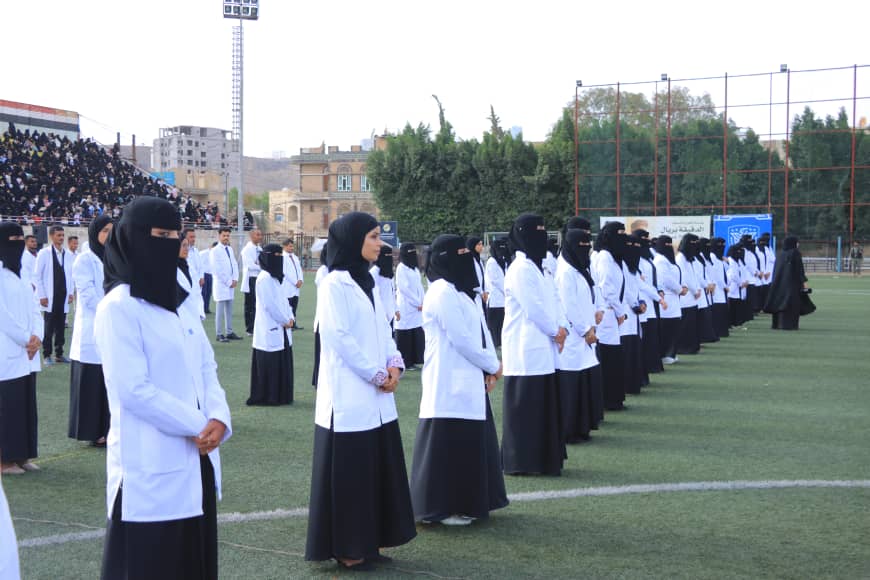 1003982284 - جامعة 21 سبتمبر Rows of women in white coats and black hijabs stand in neat rows on a stadium field during a ceremony, with spectators in the stands behind them.