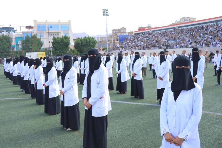 1003982287 - جامعة 21 سبتمبر Group of women in white lab coats and black niqabs standing in formation on a sports field during a ceremony.