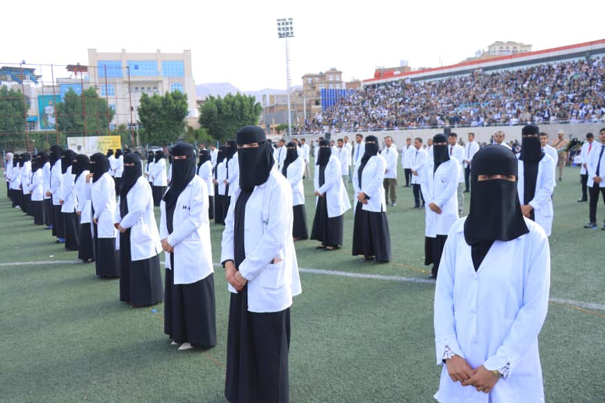 1003982290 - جامعة 21 سبتمبر Women in white lab coats and black niqabs stand in orderly rows on a sports field, with a large crowd seated in the stadium stands behind them.