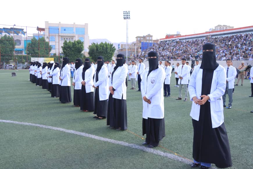 1003982293 - جامعة 21 سبتمبر Line of women in white lab coats and black abayas with niqabs standing on a sports field during a ceremony, with a large crowd in the stands behind.