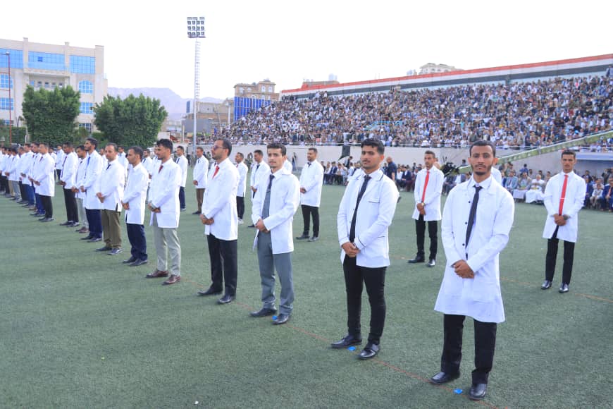 1003982296 - جامعة 21 سبتمبر Medical students in white coats stand in a line on a sports field during a ceremony, with spectators in the stands behind them.