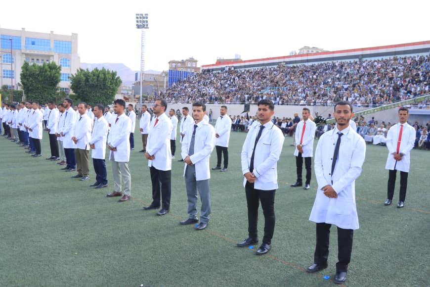 1003982299 - جامعة 21 سبتمبر Line of men in white lab coats and business attire standing in rows on a stadium field during a ceremony, with a large crowd in the stands behind them.