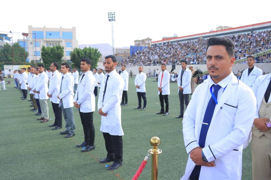 1003982302 - جامعة 21 سبتمبر Line of men in white lab coats standing at attention on a sports field during a formal ceremony, with spectators in the bleachers behind them.