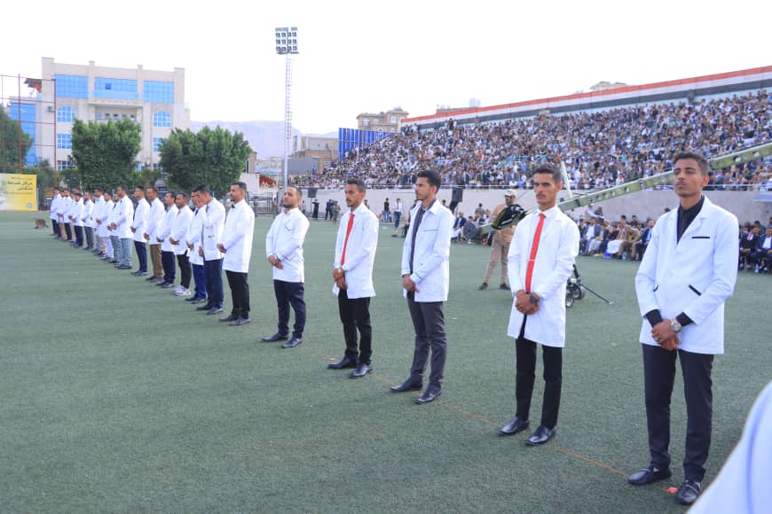 1003982305 - جامعة 21 سبتمبر Line of students in white coats standing at attention on a football field during a ceremony, with a large crowd in the stands behind them.