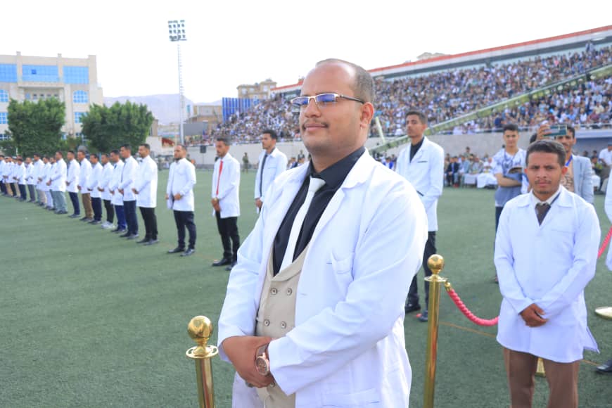 1003982308 - جامعة 21 سبتمبر Man in a white lab coat and beige vest stands with folded hands at a formal ceremony on a sports field, with spectators in the stands behind him.