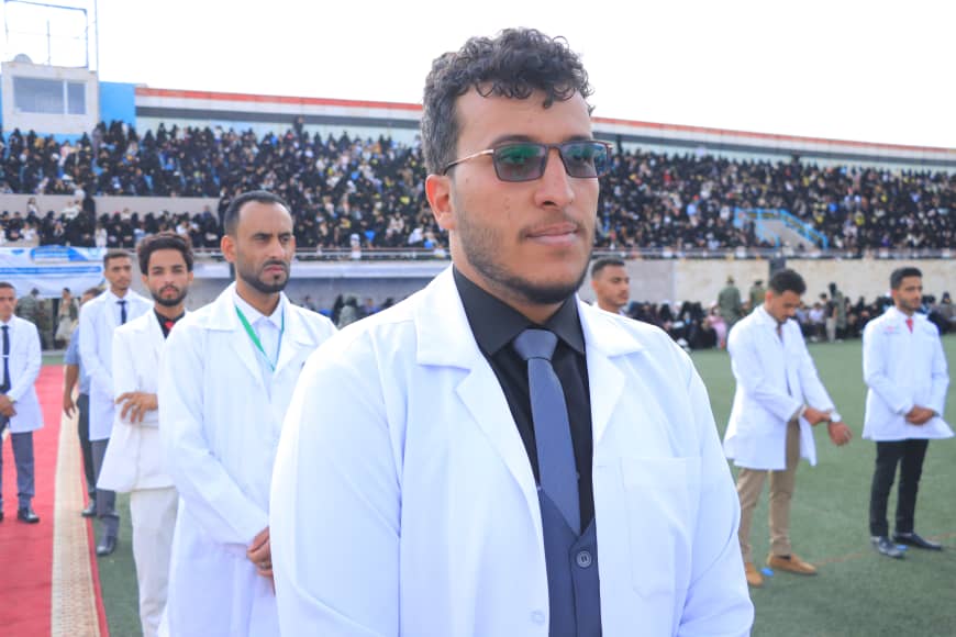1003982311 - جامعة 21 سبتمبر Young man in a white lab coat and dark tie at a field ceremony, with a crowd in bleachers behind him.