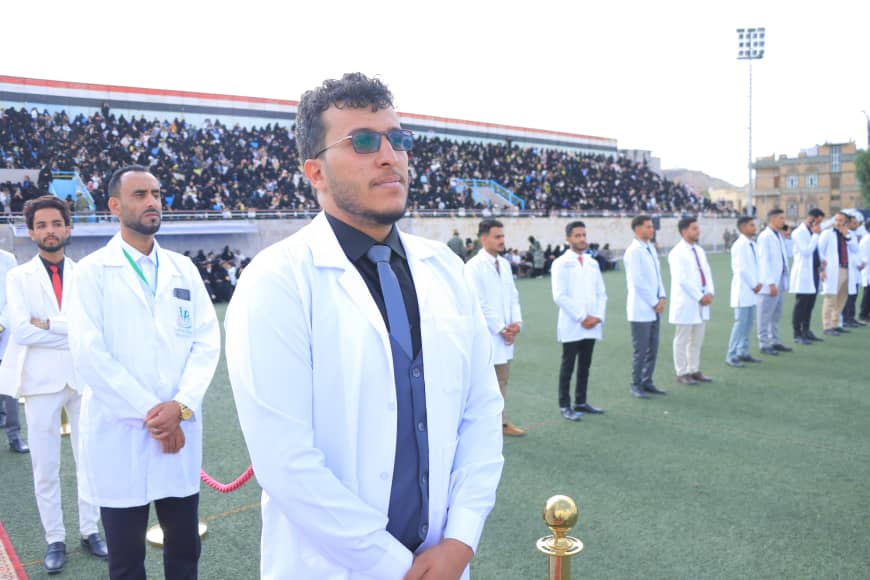 1003982314 - جامعة 21 سبتمبر Medical students in white coats stand in a line on a sports field during a ceremony, with a large audience in the stands behind them.