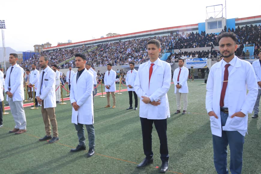 1003982317 - جامعة 21 سبتمبر Group of men in white lab coats standing in a line on a stadium field during a ceremony, with a large crowd in the stands.