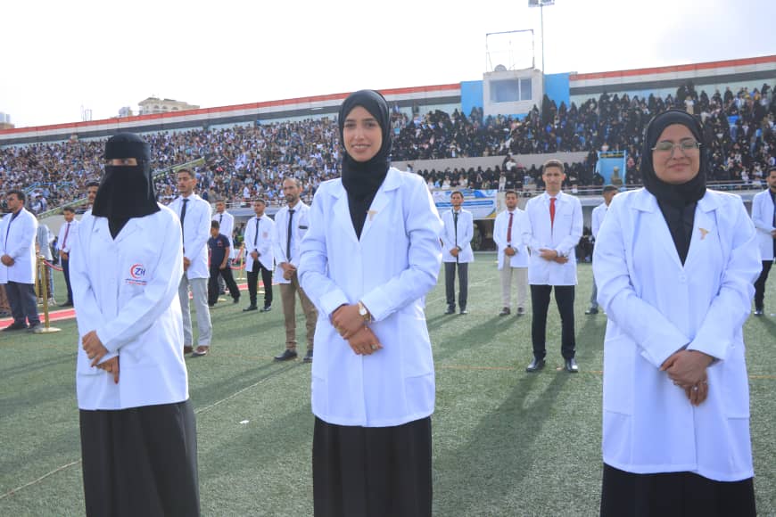 1003982320 - جامعة 21 سبتمبر Three women in white lab coats and hijabs stand on a football field during a ceremony, with a crowded stadium behind them.