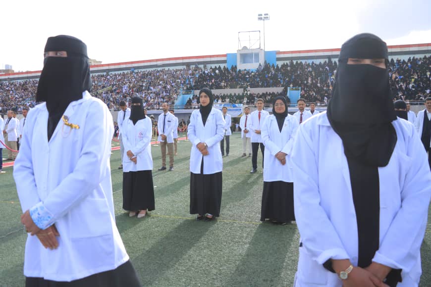 1003982323 - جامعة 21 سبتمبر Group of women in white lab coats and black hijabs standing in formation on a football field as a large crowd watches from the stands.