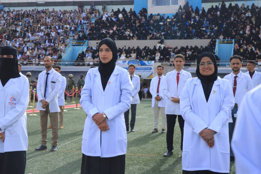 1003982326 - جامعة 21 سبتمبر Medical students in white coats stand on a sports field during a ceremony, with a large audience in the bleachers behind them, including women in hijabs in front center.