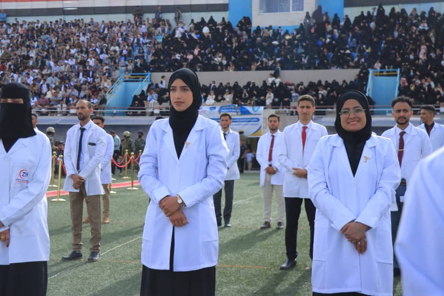 1003982329 - جامعة 21 سبتمبر Female medical students in white coats stand in a line on a sports field during a ceremony, with a large crowd watching from the bleachers behind them.