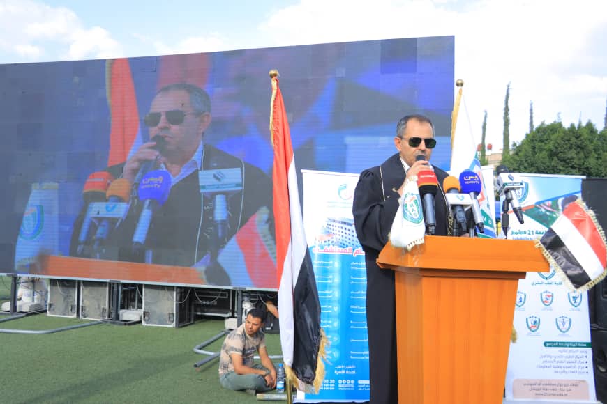1003982332 - جامعة 21 سبتمبر Man in a suit and sunglasses speaks at a wooden podium with multiple microphones at an outdoor event, flags nearby and a large screen behind him.