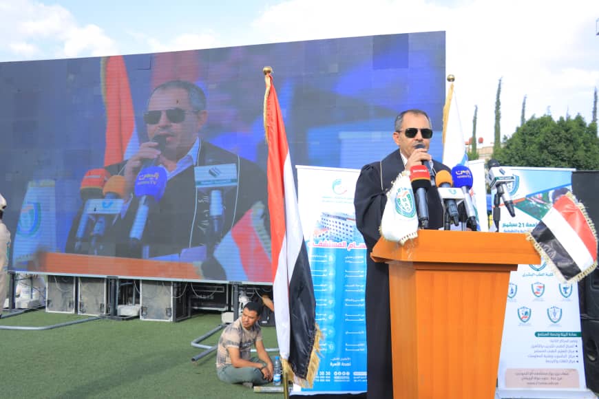 1003982335 - جامعة 21 سبتمبر Man in suit speaking at a wooden podium with multiple microphones, outdoor event with flags and a large screen behind him.