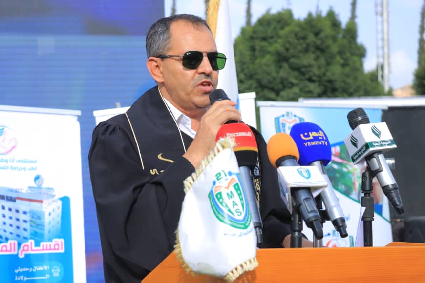 1003982338 - جامعة 21 سبتمبر Man in sunglasses speaks into a microphone at a press conference, standing behind a podium with multiple news microphones.
