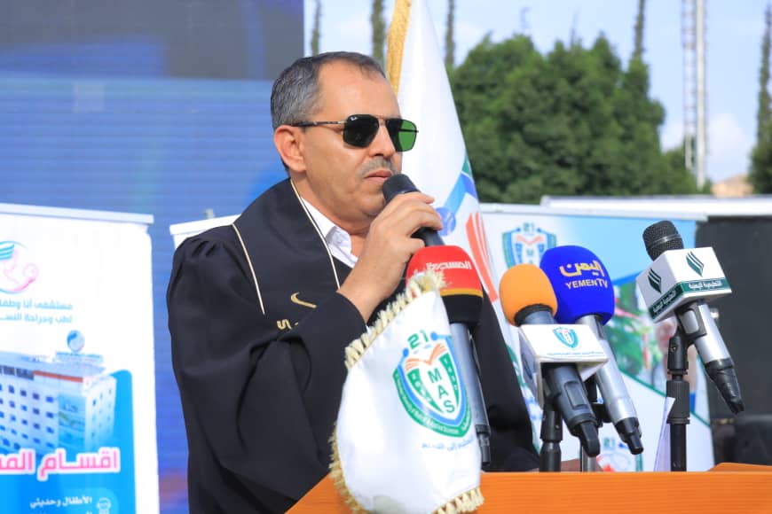1003982341 - جامعة 21 سبتمبر Man in a dark jacket wearing sunglasses speaks into a microphone at a press podium, with multiple reporters' microphones in front of him and flags in the background.