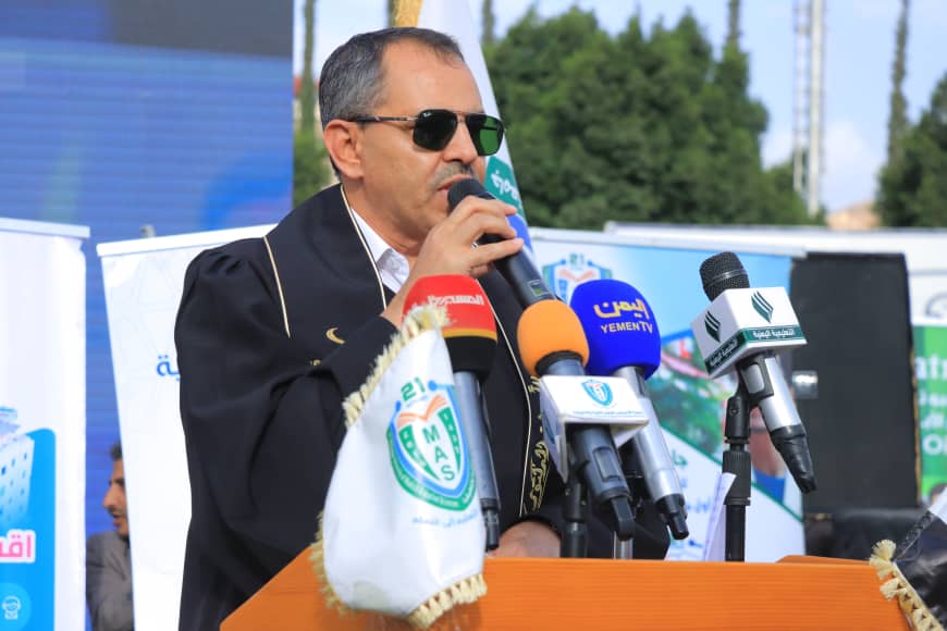 1003982344 - جامعة 21 سبتمبر Man in a dark suit and sunglasses speaks into a microphone at a podium during an outdoor press conference, with several microphones in front of him.