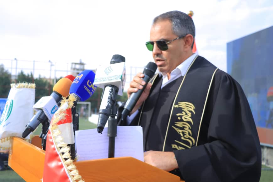 1003982347 - جامعة 21 سبتمبر Man in a black ceremonial robe and sunglasses speaks into a microphone at a podium with multiple news microphones in front of him outdoors.