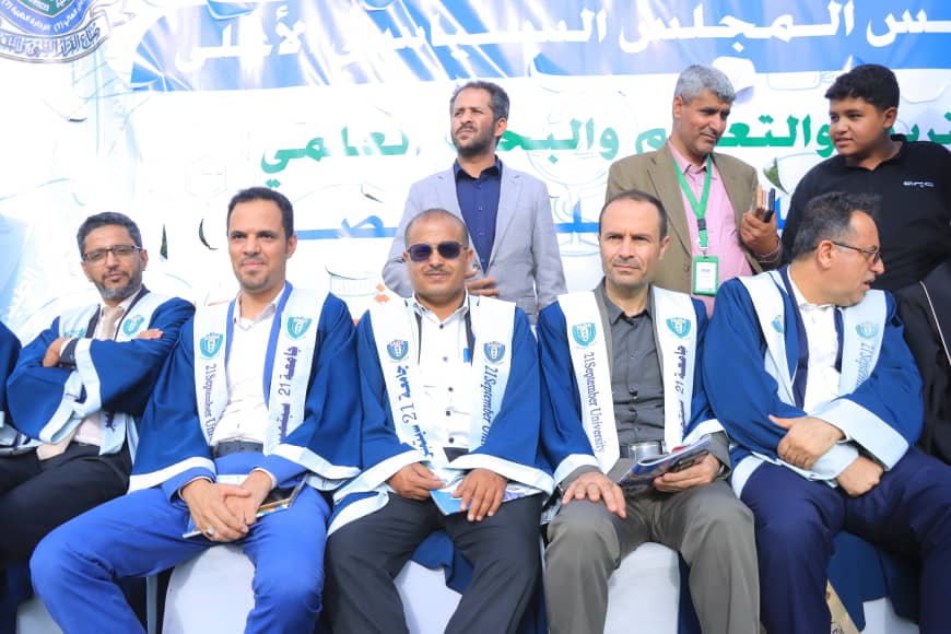1003982350 - جامعة 21 سبتمبر Group of men in blue and white graduation robes seated on stage during an Arabic-language ceremony, with a banner in the background.