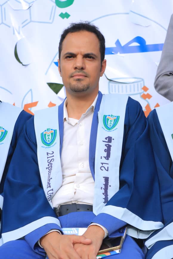 1003982355 - جامعة 21 سبتمبر A man in a blue graduation gown sitting with hands clasped, ceremonial sash displaying a crest and text, indoor backdrop behind him.