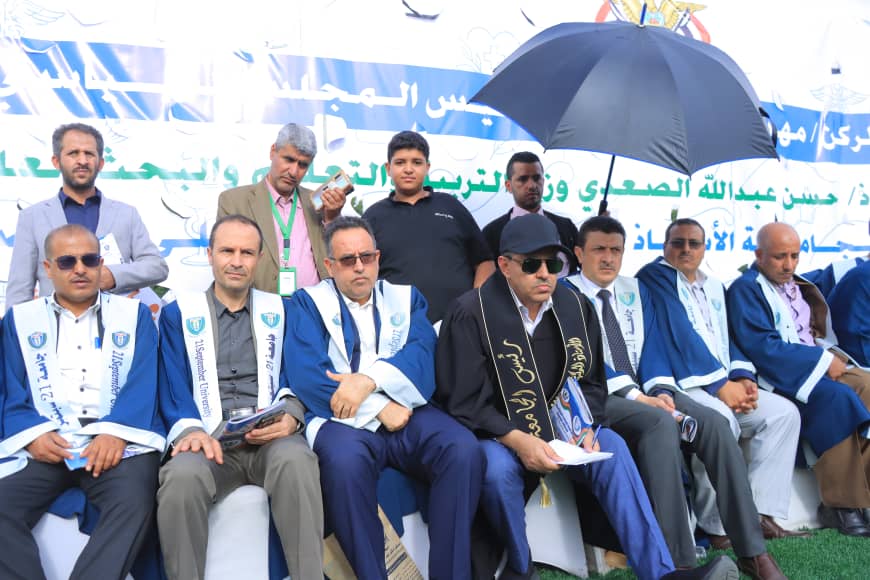 1003982358 - جامعة 21 سبتمبر Group of men in blue ceremonial robes and sashes seated at an outdoor event, a banner with Arabic text in the background.