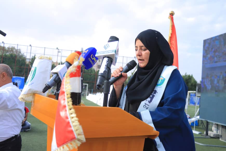 1003982361 - جامعة 21 سبتمبر Woman in a hijab speaks into a microphone at a podium during an outdoor press event, with multiple mic flags nearby.
