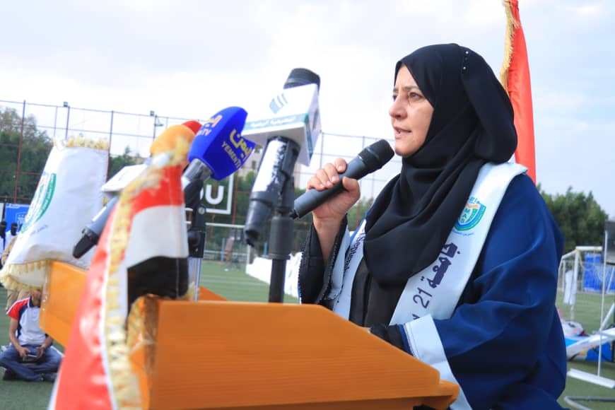 1003982364 - جامعة 21 سبتمبر Woman in a black hijab speaks into a microphone at a wooden podium on a sports field, with banners and microphones nearby.