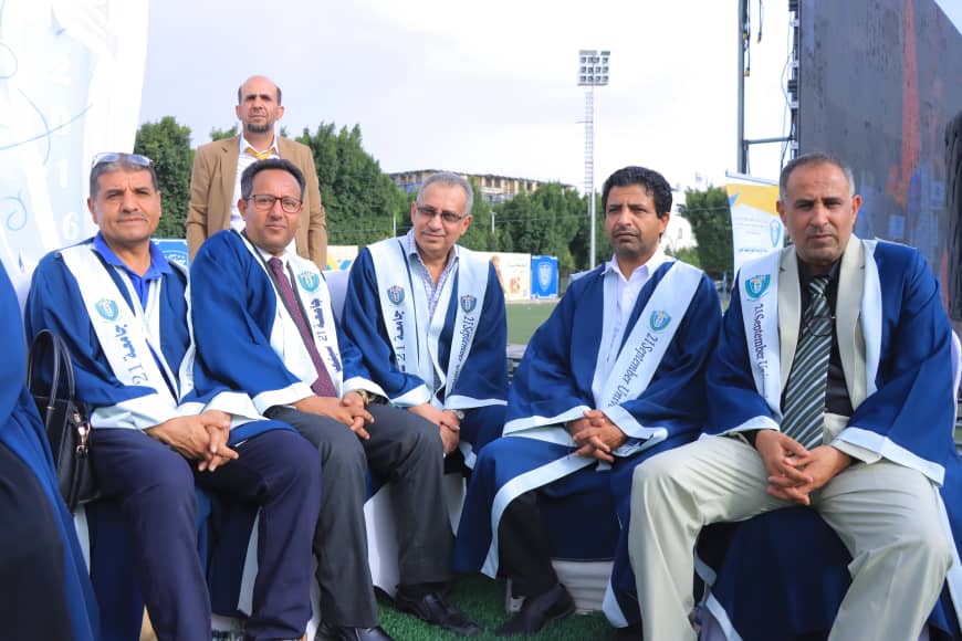 1003982367 - جامعة 21 سبتمبر Six men in blue and white ceremonial robes sit together on a bench at an outdoor stadium field with banners nearby.