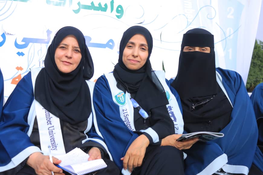 1003982370 - جامعة 21 سبتمبر Three women in blue graduation robes sit together at an event, two wearing hijabs and one wearing a niqab, smiling at the camera.