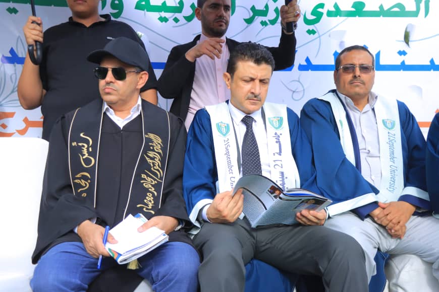 1003982373 - جامعة 21 سبتمبر Three men in blue and black graduation robes sit on a stage, reading booklets, with a banner in Arabic behind them.