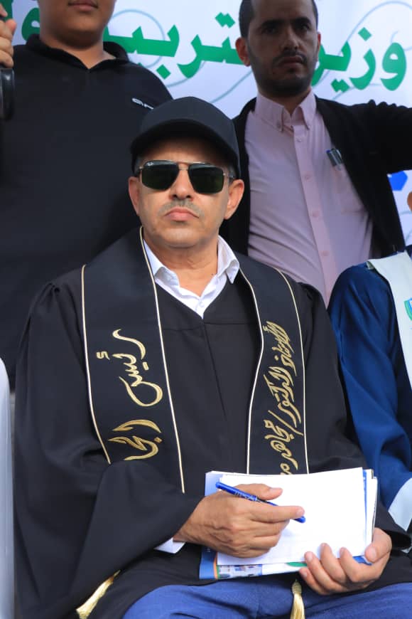 1003982376 - جامعة 21 سبتمبر Man in a black graduation gown and cap, wearing sunglasses, seated and holding papers with a blue pen at an outdoor ceremony.