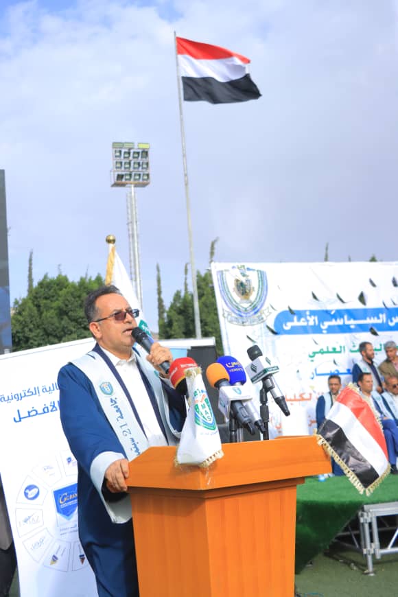 1003982379 - جامعة 21 سبتمبر Man in a blue suit and white sash speaks into a microphone at a wooden podium with several news microphones, outdoors, with banners and a tricolor flag in the background.