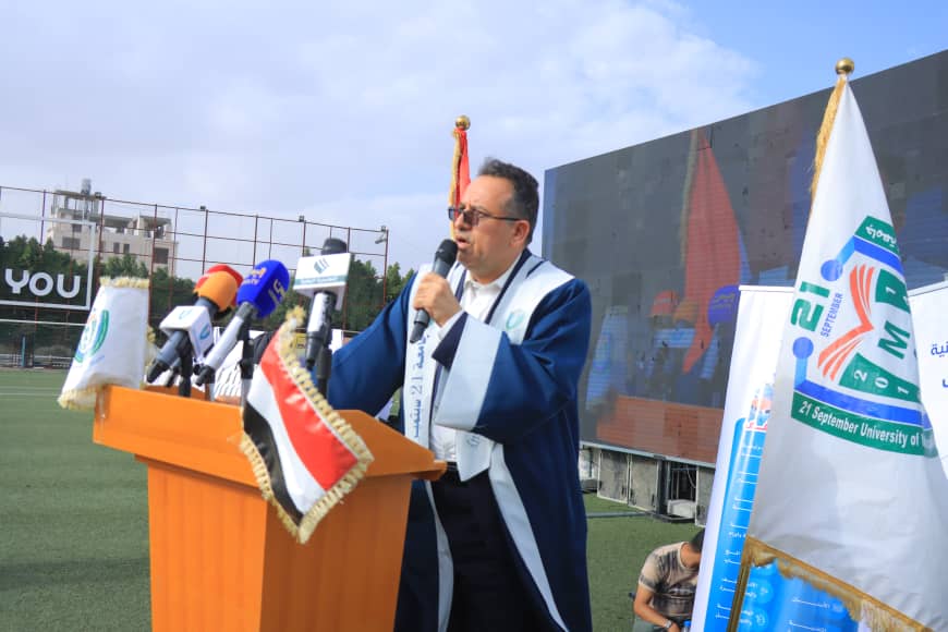 1003982382 - جامعة 21 سبتمبر Man in a blue ceremonial gown speaks at a podium with microphones during an outdoor university event, flags in the background.