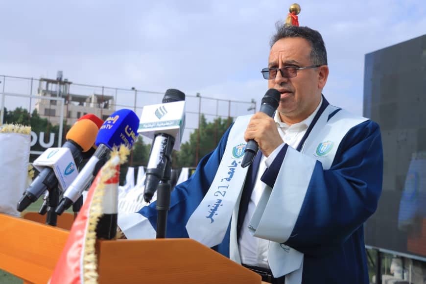 1003982385 - جامعة 21 سبتمبر Man in blue ceremonial robe and sash speaks into a handheld microphone at a podium with multiple news microphones on an outdoor stage
