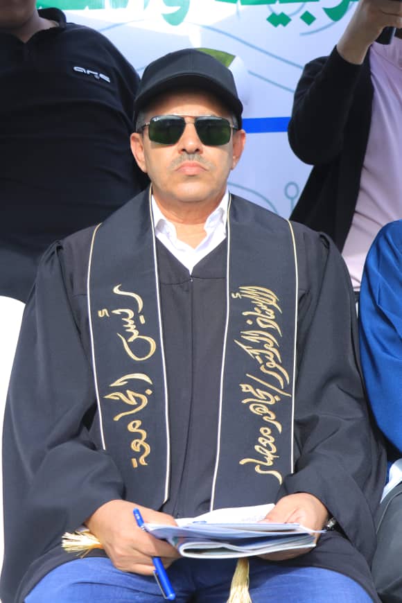 1003982388 - جامعة 21 سبتمبر Man wearing a black graduation gown and cap with gold embroidery, sunglasses, and a white shirt, sitting and holding papers and a blue pen outdoors.