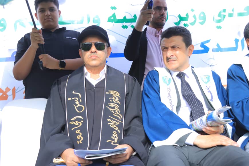 1003982391 - جامعة 21 سبتمبر Two men in ceremonial robes seated on a stage during a formal event; one wears a black robe with gold script and sunglasses, the other a blue robe with a sash, holding a rolled document, with a banner in the background.